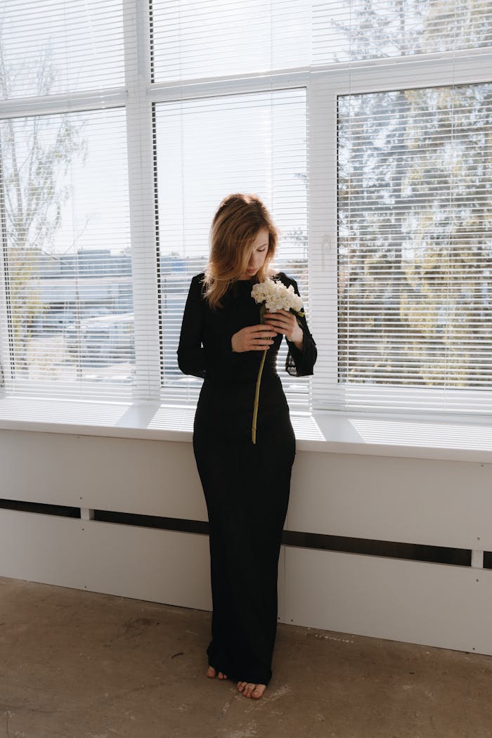 creative-03 Elegant woman in black dress standing by a window with white flowers.
